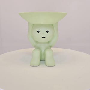 May include: A light green figurine of a person holding a shallow dish above their head. The figurine has a minimalist design with black eyes and a small mouth. It sits on a white, round surface.