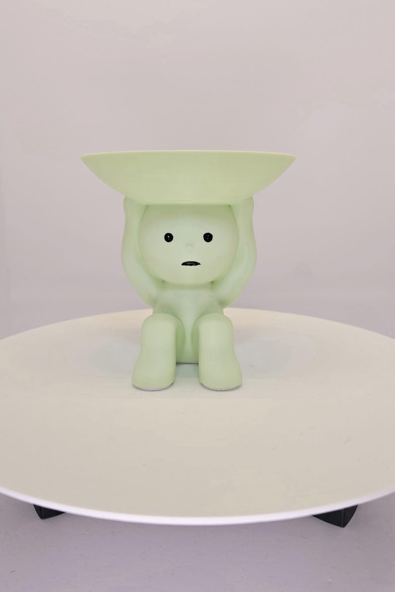 May include: A light green figurine of a person holding a shallow dish above their head. The figurine has a minimalist design with black eyes and a small mouth. It sits on a white, round surface.