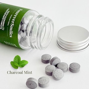 Op de afbeelding: Een heldere glazen fles met een zilveren deksel, met grijze houtskool-munt-tandpastatabletten. De fles is gelabeld met "FourEverSmiles" met groene tekst. Een takje munt en de woorden "Charcoal Mint" zijn ook zichtbaar.