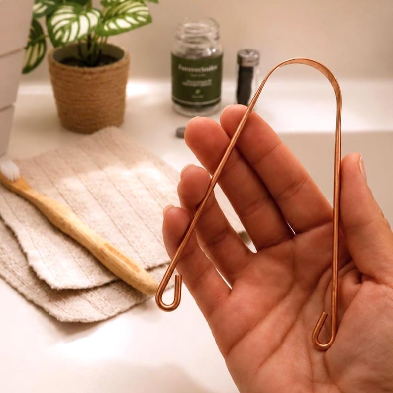 Hand holding a copper tongue scraper above a bathroom sink with neutral decor, bamboo toothbrush, and calming wellness-inspired styling.