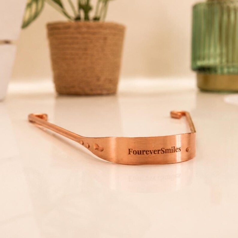 Copper tongue scraper resting on a clean white surface with soft natural lighting, featuring a minimal aesthetic oral care setup by FoureverSmiles.