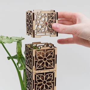 May include: Wooden plant pot with a decorative floral design. The pot is made of light brown wood and has an open, cage-like structure. The pot is shown stacked with another pot, and a hand is holding a third pot. The pots are filled with soil and a small plant.