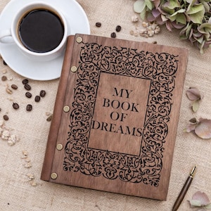 May include: A wooden journal with the text "MY BOOK OF DREAMS" in a decorative frame. The journal is open. A cup of coffee and coffee beans are next to the journal.