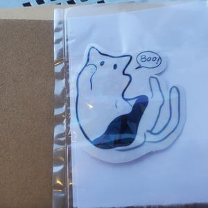 Könnte beinhalten: Eine weiße Geisterkatze mit einem schwarzen Schwanz und einer Sprechblase, die "Boo!" auf weißem Hintergrund sagt.
