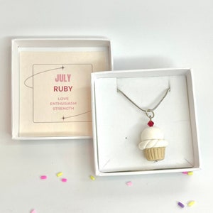 May include: A white cupcake charm necklace with a red gem on top, in a white box with a card that says "JULY RUBY LOVE ENTHUSIASM STRENGTH"