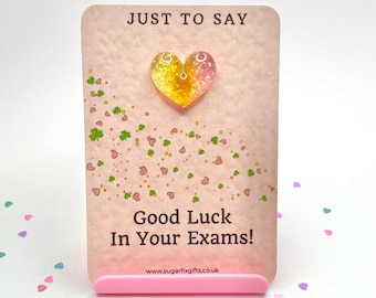 May include: A pink card with the text "Just to Say" and "Good Luck In Your Exams!" with a small yellow and pink heart charm. The card is decorated with small green shamrocks and pink hearts.