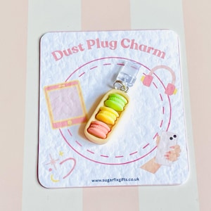 May include: A decorative dust plug charm with three miniature macarons in pink, yellow, and green, set in a light brown holder. The charm is attached to a clear plastic plug. The background is a card with the text "Dust Plug Charm".