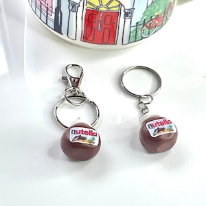 Pode incluir: Dois chaveiros prateados com um charme de pote de Nutella marrom e branco. Os charms são redondos e têm o logotipo da Nutella.