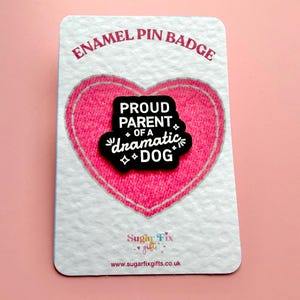 Pode incluir: Um distintivo de esmalte num cartão branco com um desenho de coração rosa. O pin preto diz "PROUD PARENT of a dramatic DOG" em texto branco. O cartão tem o texto "ENAMEL PIN BADGE" e o endereço do site.