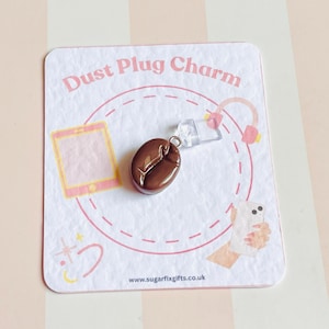 Peut inclure: Un charm de prise anti-poussière en forme de grain de café marron sur une carte blanche avec le texte "Dust Plug Charm". La carte a des accents roses et des illustrations.
