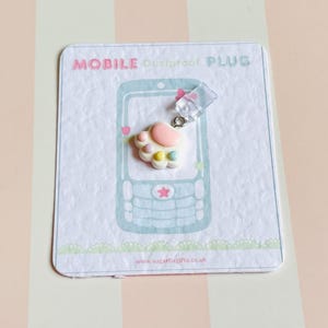 Peut inclure: Un bouchon anti-poussière pour mobile avec un motif de patte de couleur pastel. Le bouchon est fixé à un morceau de plastique transparent et se trouve sur une carte avec une illustration de téléphone et le texte "MOBILE Dustproof PLUG".