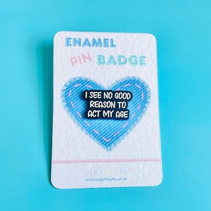 May include: An enamel pin badge on a white card with a blue heart design. The pin reads "I SEE NO GOOD REASON TO ACT MY AGE" in white text on a black background. The card has the words "ENAMEL PIN BADGE" printed on it.