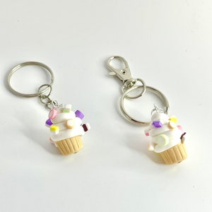 Pode incluir: Dois chaveiros prateados com pingentes de cupcake branco. Os cupcakes são decorados com doces e granulados coloridos.
