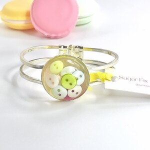 May include: A silver bangle bracelet with a clear round charm filled with colourful buttons. The charm is surrounded by a silver rim. The bracelet is on a white surface with a yellow ribbon and a white tag that says "Sugar Fix @junkee"