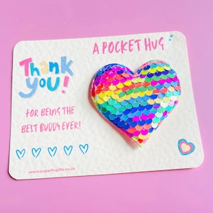 Puede incluir: Una tarjeta "Pocket Hug" con un corazón de lentejuelas. La tarjeta dice "¡Gracias! Por ser el mejor amigo del mundo!" en texto colorido. El corazón está hecho de lentejuelas de colores del arcoíris. El fondo es rosa.