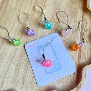 May include: A collection of colorful phone charms with a mushroom design. The charms are in various colors, including green, purple, pink, orange, and blue. Each charm has a silver loop and is attached to a small card with a phone graphic.