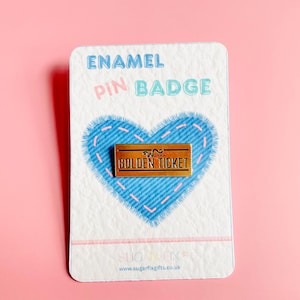 Peut inclure: Un badge en émail sur une carte blanche avec un motif de cœur bleu. L'épingle est de forme rectangulaire avec les mots "GOLDEN TICKET" en lettres noires. La carte porte également les mots "ENAMEL PIN BADGE" en bleu et rose.