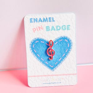 May include: A red treble clef enamel pin badge on a white card with a blue denim heart design. The card has the words "ENAMEL PIN BADGE" in light blue and pink lettering. The background is a soft pink.