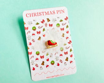 May include: A Christmas pin on a white card with a festive pattern. The pin is a red ice skate with green and gold accents. The card reads "CHRISTMAS PIN" in red text and has a light blue background.
