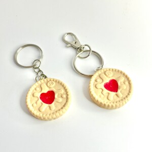 May include: Two keyrings with biscuit-shaped charms, each with a red heart centre. The charms are cream-coloured with a decorative edge. One keyring has a split ring, the other a lobster clasp.