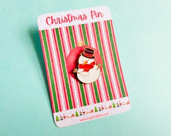 May include: A Christmas pin depicting a snowman wearing a black top hat and a red scarf. The pin is displayed on a card with a red and green striped design and the text "Christmas Pin". The background is a light blue colour.