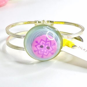 Può includere: Un braccialetto rigido argentato con un bottone rosa e viola con un disegno a fiocco di neve. Il bottone è racchiuso in una cupola di vetro trasparente.
