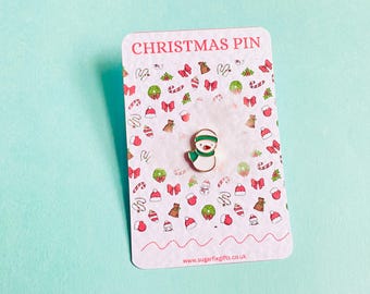 May include: A Christmas pin with a snowman design, white body, green scarf and hat. The pin is on a card printed with "CHRISTMAS PIN" and a pattern of festive illustrations. The background is a light blue colour.