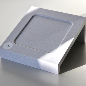 Könnte beinhalten: Ein hellgrauer, abgewinkelter Präsentationsständer mit einem eingelassenen quadratischen Bereich. Auf der Vorderseite befindet sich ein kleiner, runder Aufkleber. Der Ständer besteht aus einem strukturierten Material und wird von einer starken Lichtquelle beleuchtet.