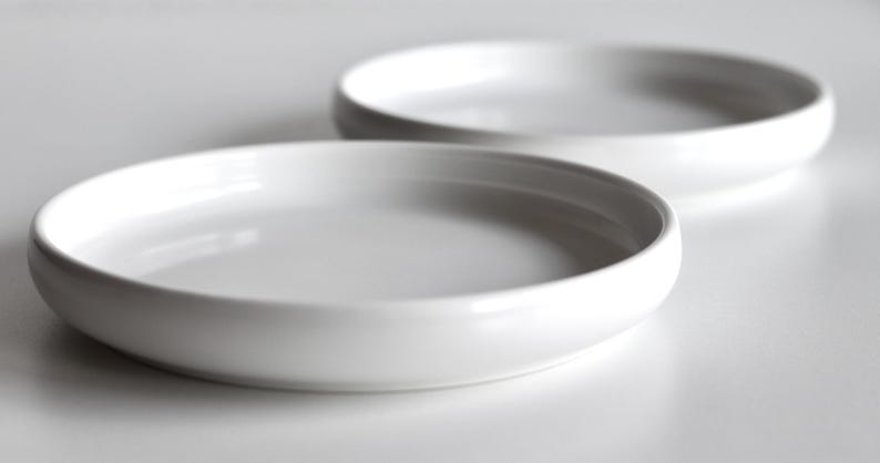 May include: Two white ceramic plates, one stacked on top of the other. The plates are round and have a slightly raised rim.