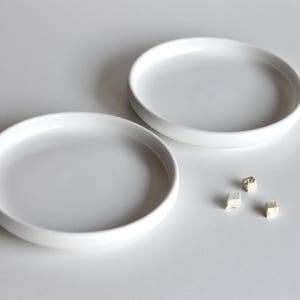 May include: Two white ceramic dishes, one slightly larger than the other, are arranged on a white surface. Three small, white, cube-shaped objects are scattered near the smaller dish.