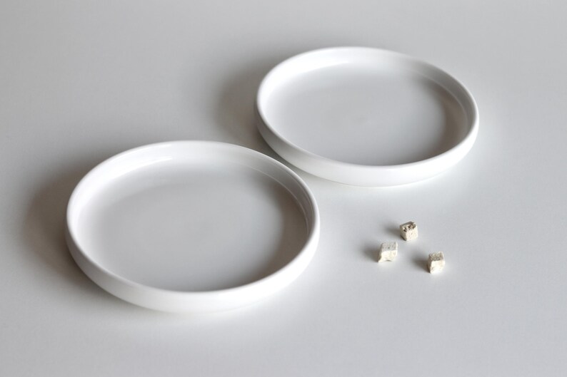 May include: Two white ceramic dishes, one slightly larger than the other, are arranged on a white surface. Three small, white, cube-shaped objects are scattered near the smaller dish.