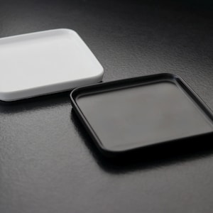 Op de afbeelding: Twee vierkante keramische trays, één wit en één zwart, worden getoond op een donkere ondergrond. De trays hebben afgeronde hoeken en een minimalistisch ontwerp, geschikt voor serveren of presenteren.