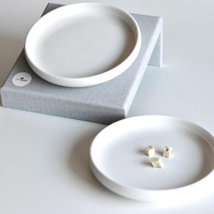 May include: Two white ceramic dishes, one on top of the other, on a gray platform. The top dish is larger than the bottom dish. The bottom dish has three small, white, geometric shapes on it.