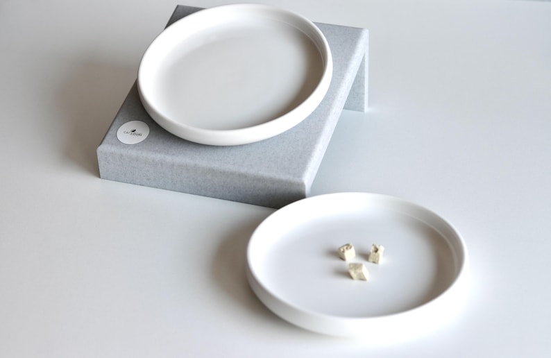 May include: Two white ceramic dishes, one on top of the other, on a gray platform. The top dish is larger than the bottom dish. The bottom dish has three small, white, geometric shapes on it.