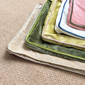 May include: A stack of five rectangular velvet pillow covers in various colors, including beige, olive green, chartreuse, white, and pink. Each cover features a contrasting piped edge in a different color, creating a layered, textured appearance.