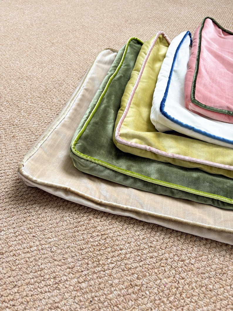 May include: A stack of five rectangular velvet pillow covers in various colors, including beige, olive green, chartreuse, white, and pink. Each cover features a contrasting piped edge in a different color, creating a layered, textured appearance.