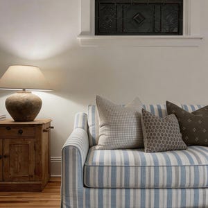 Op de afbeelding: Een woonkamer met een blauw-wit gestreepte bank en decoratieve kussens. Een houten bijzettafel met een lamp en een glas-in-loodraam zijn ook zichtbaar. De kamer heeft een neutrale kleurstelling.