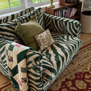 Könnte beinhalten: Ein gemütliches Wohnzimmer mit einem Sofa mit Zebra-Streifen in Grün und Creme. Das Sofa ist mit olivgrünen und floralen Kissen sowie einer Patchwork-Steppdecke ausgestattet. Ein gemusterter Teppich und ein Bücherregal aus Holz ergänzen das Ambiente.
