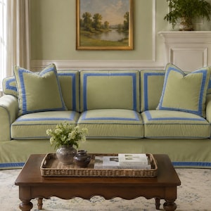 May include: A three-cushion sofa upholstered in light green fabric with blue trim accents along the edges and seams. The sofa features rolled arms, a skirted base, and two matching square throw pillows with blue borders.