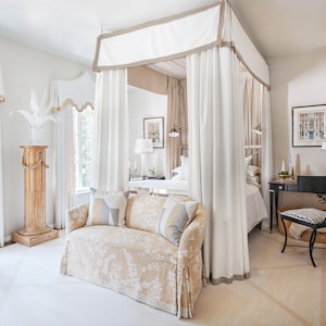 Puede incluir: Dormitorio luminoso con cama con dosel y cortinas blancas. Un sofá con estampado floral y cojines decorativos se encuentra frente a la cama. La habitación cuenta con una columna de madera, un escritorio negro y una silla con estampado de cebra.