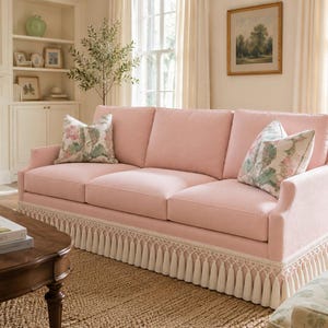 May include: A three-cushion sofa upholstered in light pink fabric, featuring a decorative cream-colored fringe trim along the base. The sofa includes two square throw pillows with a floral pattern in shades of green, pink, and white.