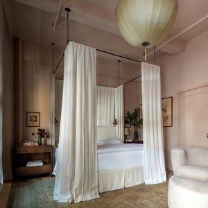 May include: A bedroom with a four-poster bed draped with white curtains. The room has a soft, pinkish-beige color scheme, with a large, round light fixture hanging from the ceiling. A small wooden nightstand and a white armchair are also visible.