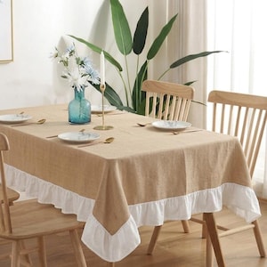 May include: A beige rectangular tablecloth with a white ruffled edge, set on a wooden table. The table is set with plates, spoons, and a blue glass vase with flowers. Wooden chairs surround the table, creating a dining setting.