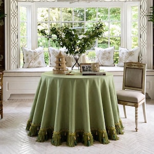 May include: A round, light green tablecloth with a decorative fringe. The table is set with a small pagoda, a vase of greenery, and a framed photo. A chair with a light-colored seat and a decorative back is next to the table.