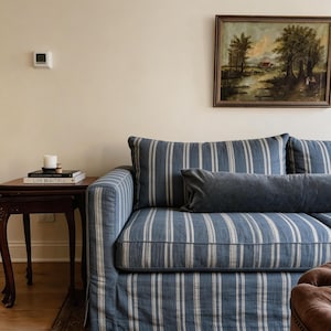 May include: A blue and white striped sofa with a matching skirt and two blue pillows. A small wooden side table with books and a candle sits to the left of the sofa. A framed landscape painting hangs on the wall.