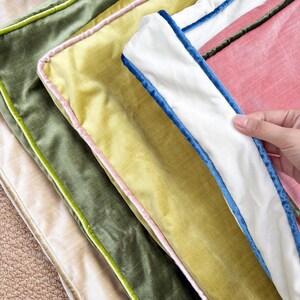 May include: A stack of four rectangular velvet cushion covers featuring contrasting piping. The colors include a beige base, an olive green cover with lime piping, a yellow cover with pink piping, and a pink cover with a white border and blue piping.