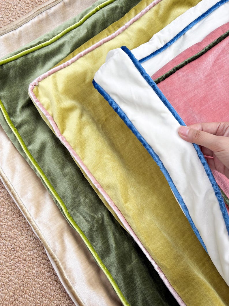 May include: A stack of four rectangular velvet cushion covers featuring contrasting piping. The colors include a beige base, an olive green cover with lime piping, a yellow cover with pink piping, and a pink cover with a white border and blue piping.