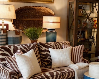 May include: A living room scene featuring a zebra-print sofa with white throw pillows. A large woven basket and framed artwork hang on the wall. Lamps with striped shades and a blue base provide lighting. Decorative items and a cabinet complete the cozy interior.