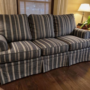 May include: A three-cushion sofa with a blue and white striped pattern. The sofa has rolled arms and a skirted base. The cushions and skirt have a vertical stripe design. The sofa is in a room with a wooden floor and a window.