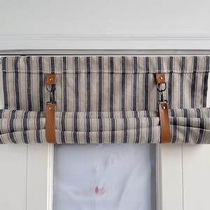 Op de afbeelding: Een gestreepte rolgordijn met bruine leren banden en metalen ringen. Het rolgordijn is gemonteerd op een wit raamkozijn.